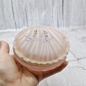 Può includere: Una scatola portaoggetti in vetro rosa a forma di conchiglia con un design floreale. Il design presenta fiori viola e foglie verdi.