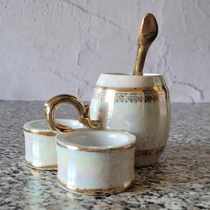 May include: A white porcelain salt and pepper shaker set with a gold trim and a spoon. The set has a pearlescent finish and is decorated with a gold band.