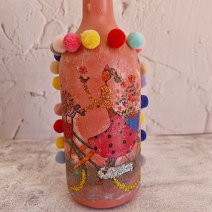 Upcyled wine bottle painted in pink and decoupaged witha picture of a girl riding a bile. There are also decorative colorful pompons.