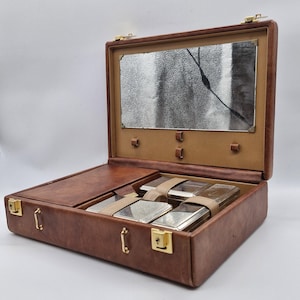 May include: Open brown leather travel case with a mirror and several silver containers secured with beige straps. The case has gold-coloured latches and hinges, and a beige interior.