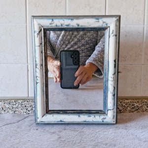 Può includere: Uno smartphone nero con un obiettivo della fotocamera sul retro si riflette in uno specchio incorniciato di bianco e blu.