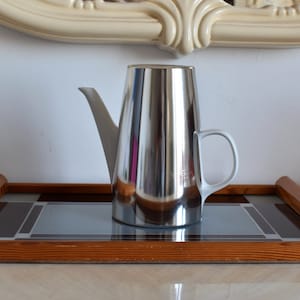 Può includere: Una caffettiera in metallo argentato con manico bianco su un vassoio di legno con un motivo geometrico.