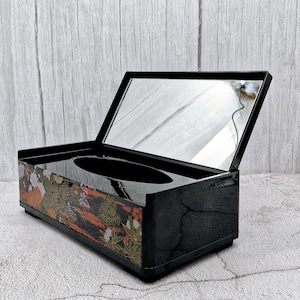 May include: Black tissue box with a floral design and a mirror lid. The lid is open and the tissue box is sitting on a gray surface.