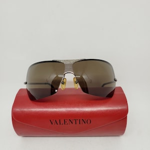 以下が含まれることがあります： シルバーフレームのブラウンのサングラスと、"VALENTINO"と印刷された赤いケース。