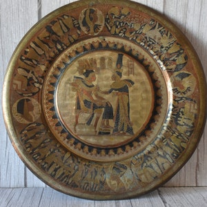 Könnte beinhalten: Ein runder Messingteller mit einem komplizierten Design, das eine ägyptische Szene mit zwei Figuren in Gold und Schwarz zeigt. Der Teller hat einen schwarzen und goldenen Rand mit einem sich wiederholenden Muster.