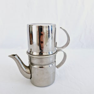 Puede incluir: Una cafetera de acero inoxidable plateada con un filtro extraíble y un asa. La cafetera está hecha de dos piezas apiladas.