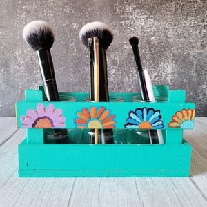 May include: A turquoise wooden crate with four glass jars for holding makeup brushes. The jars are decorated with colorful flowers. The crate is sitting on a white wooden surface.
