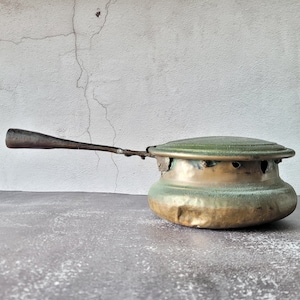 Pode incluir: Uma chaleira vintage de latão e metal verde com uma alça longa. A chaleira tem uma pátina desgastada e envelhecida e é projetada para ser usada em um fogão.