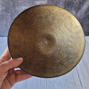May include: A large, round, hammered brass bowl with a slightly raised center. The bowl has a warm, golden color and a textured surface.