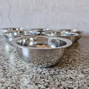 Peut inclure: Un ensemble de sept bols à mélanger en acier inoxydable. Les bols sont ronds avec une surface brillante et réfléchissante. Ils sont disposés sur un comptoir moucheté gris et marron, avec un mur gris clair en arrière-plan.