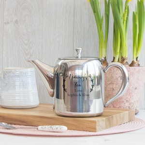 May include: A polished stainless steel teapot with a curved spout and handle. The side features a black engraved design of a laurel wreath surrounding the text Nanny's Tea, Love, Sophia & Elise. The teapot has a lid with a small knob.