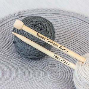 May include: Two wooden knitting needles with the text "Happy Birthday Nanny" and "Love Elise x" are laid across two balls of yarn. One ball is gray, and the other is off-white. The needles are on a gray, textured surface.