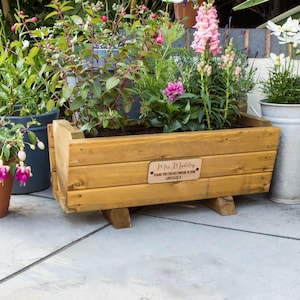 May include: A wooden planter box, filled with a variety of plants and flowers. The box is light brown and rests on two wooden supports. A small wooden plaque on the front reads "Mrs. Madeley, THANK YOU FOR HELPING ME BLOOM LOVE ELISE Y."