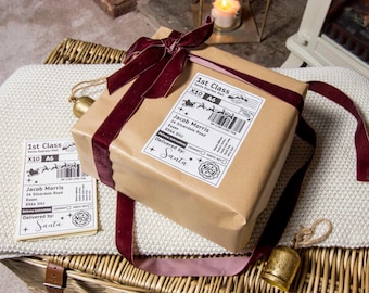 May include: A wrapped gift in brown paper with a burgundy velvet ribbon and bow. The gift has a shipping label that reads "1st Class" and "Delivered by: Santa". A small gold bell and a candle in a lantern are also visible.