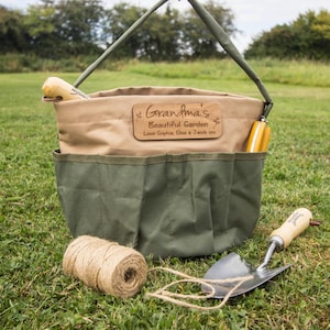 May include: A green and beige canvas garden tool bag with a handle, pockets, and a wooden plaque that reads "Grandma's Beautiful Garden". A ball of twine and a trowel are placed on the grass next to the bag.