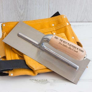 May include: A personalised trowel with a wooden handle engraved with the message "Dad - The Best Plasterer In Town Love Sophia, Elise & Jacob". The trowel is resting on a yellow leather tool belt.