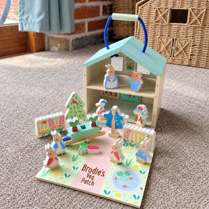 May include: A wooden toy house with a light blue roof and a blue handle, featuring multiple animal figurines, a wheelbarrow, a tree, and fences. The base is printed with the text Brodie's Veg Patch and includes a small pond illustration.