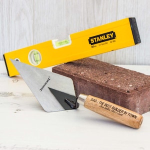 May include: A wooden-handled trowel with a metal blade and the words "DAD: THE BEST BUILDER IN TOWN" engraved on the handle. A yellow Stanley level and a red brick are also in the image.