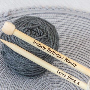 May include: A skein of gray yarn with two wooden knitting needles. The needles are engraved with the text "Happy Birthday Nanny" and "Love Elise x". The yarn is a dark gray color.