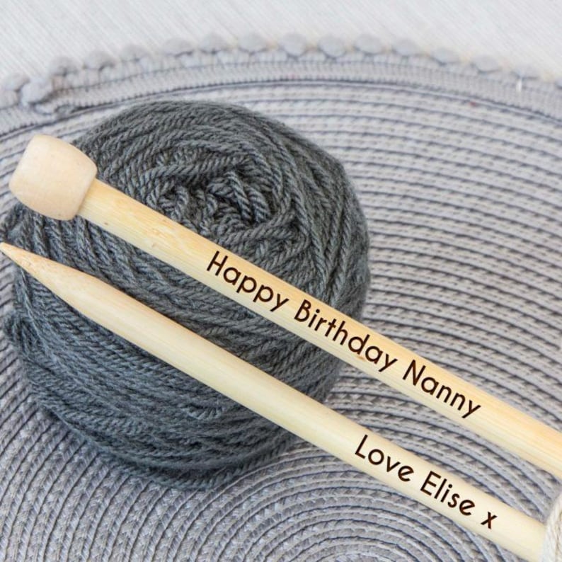 May include: A skein of gray yarn with two wooden knitting needles. The needles are engraved with the text "Happy Birthday Nanny" and "Love Elise x". The yarn is a dark gray color.