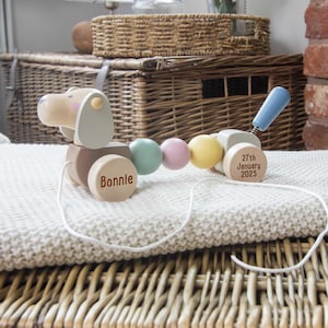 May include: A wooden pull toy shaped like a dog with a light wood finish, featuring pastel-coloured spherical beads in green, pink, and yellow. The front wheel is engraved with the name Bonnie, and the back wheel displays 27th January 2023.