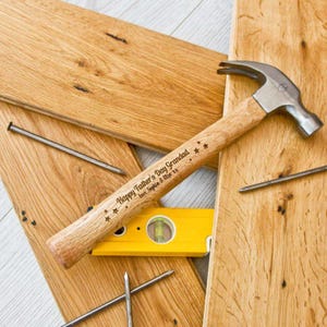May include: A wooden hammer with a black metal head and a personalised message that reads "Happy Father's Day Grandad Love Sophia & Lily xx". The hammer is lying on a wooden surface with nails and a spirit level.