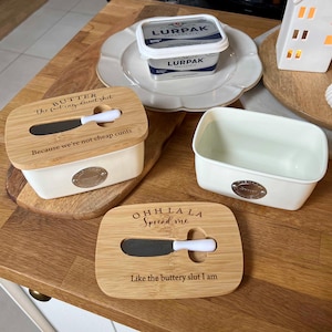 May include: Two rectangular butter dishes with cream-coloured ceramic bases and bamboo lids. Each lid features a slot for a butter knife and engraved text: one reads 'BUTTER The fact my decent shit Because we're not cheap cunts', the other 'OHH LA LA Spread me Like the buttery slut I am'.