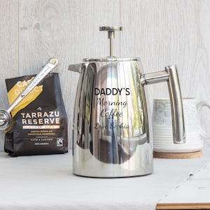 May include: A polished stainless steel French press coffee maker with a plunger lid and side handle. The body is engraved with the text DADDY'S Morning Coffee Love Sophia. A silver coffee scoop and a bag of Tarrazu Reserve coffee are nearby.