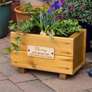 May include: A light brown wooden planter box filled with green plants and blue flowers. A wooden plaque on the front reads "Nanny, Happy Mother's Day, Love Sophia, Elise & Jacob". The planter is on small wooden feet.