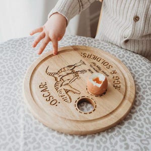 May include: A light brown wooden breakfast board with a dinosaur design and the text "OSCAR: EGG & SOLDIERS". An egg is placed on the board. The board is round and has a circular design.