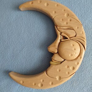 May include: Wooden crescent moon wall art with a carved face. The moon is light brown with a smiling face, detailed features, and small circular indentations. The artwork is set against a light blue background.