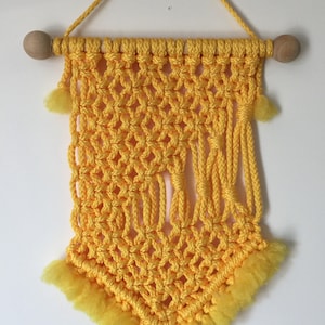 May include: A handmade yellow macrame wall hanging. The decorative piece features a woven diamond pattern, a wooden dowel, and yellow fringe. The macrame is suspended by a yellow cord and has small yellow tassels on the sides.
