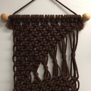 May include: A brown macrame wall hanging with a wooden dowel at the top. The macrame is made with thick brown cord and has a geometric pattern.