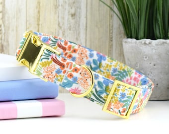 May include: A white dog collar with a floral pattern in shades of pink, orange, blue, and green. The collar has gold-colored hardware, including a buckle and a D-ring.