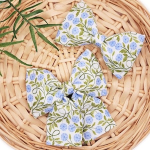 Puede incluir: Tres lazos para el pelo de tela con un estampado floral de flores azules y hojas verdes sobre un fondo blanco. Los lazos están colocados sobre una cesta de mimbre.