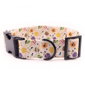 May include: A dog collar with a Halloween pattern. It has a black plastic buckle and a black metal D-ring. The beige fabric is decorated with colourful illustrations of candy apples, ghosts, and other Halloween treats.