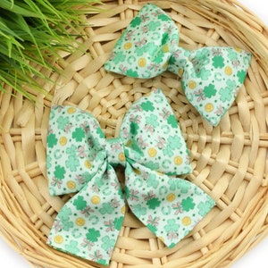 May include: Two green and white fabric hair bows with a shamrock and gold coin pattern. The bows are on a woven wicker surface.