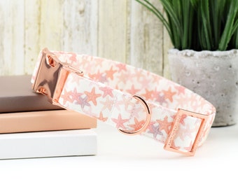 May include: A white dog collar with a pink and white starfish pattern and rose gold hardware. The collar has a buckle closure and a D-ring for attaching a leash.