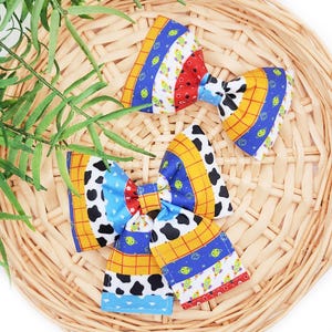 Puede incluir: Dos pajaritas de tela coloridas con patrones inspirados en una popular película de animación. Los lazos presentan diseños con detalles en rojo, azul, amarillo y negro. Están dispuestas sobre una cesta de mimbre con follaje verde.