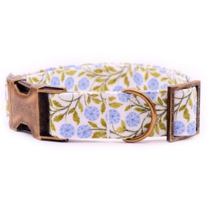 Puede incluir: Collar para perros con un estampado floral de flores azules y hojas verdes sobre fondo blanco. Hebilla y anilla en D de metal color bronce.