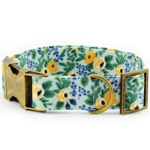 May include: A light blue dog collar with a floral pattern featuring yellow and blue flowers. The collar has a brass buckle and a brass D-ring. The text "SANDY PAWS COLLAR CO." is engraved on the buckle.
