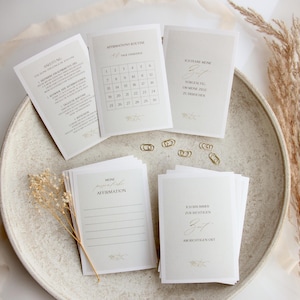 May include: A selection of white affirmation cards with gold detailing, presented on a neutral ceramic plate. The cards display printed text and a 30-day challenge grid. Dried flowers and gold paper clips add to the presentation.