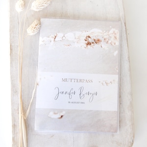 Könnte beinhalten: Eine weiße Karte mit braunem Hintergrund und dem Text "Mutterpass" in goldener Schrift. Der Name "Jennifer Berger" ist darunter in Schreibschrift geschrieben. Das Datum "19. August 1985" ist in goldener Schrift unter dem Namen gedruckt.