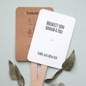 Könnte beinhalten: Ein weißer Hochzeitfächer mit dem Text "Hochzeit von Hannah & Paul" und "Schön, dass ihr da seid". Der Fächer hat ein rosa Herz und einen Holzstiel.