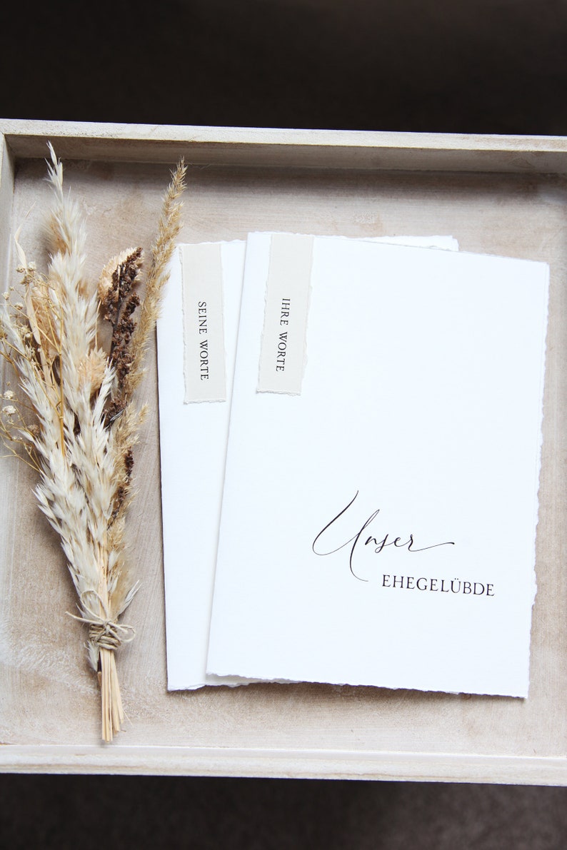 Peut inclure: Deux cartes blanches avec le texte "Ihre Worte" et "Seine Worte" dessus. Les cartes sont dans un plateau en bois avec un bouquet de fleurs s&eacute;ch&eacute;es. La plus grande carte porte le texte "Unser Ehegel&uuml;bde" en cursive.