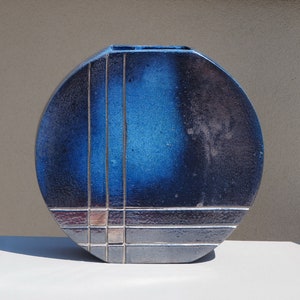 May include: A ceramic vase with a rounded, flattened shape. The vase features a gradient of blue hues, with darker shades at the edges and a lighter blue center. Silver lines and squares create a geometric pattern across the surface. The vase is a decorative home decor item.