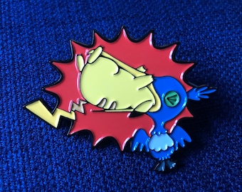 Puede incluir: Un pin de esmalte metálico que muestra a una criatura amarilla sostenida por una criatura azul similar a un pájaro sobre un fondo rojo con forma de burbuja de cómic dentada. La figura amarilla tiene una cola de rayo y la azul tiene alas.