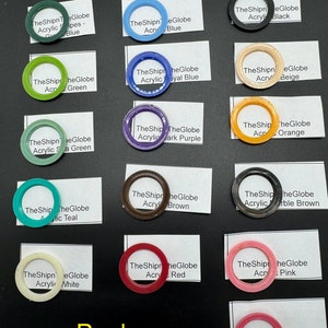 May include: A collection of fifteen circular acrylic rings in various colors including black, white, red, pink, blue, green, orange, brown, and purple. Each ring is paired with a white label identifying the color and the brand TheShipnTheGlobe.