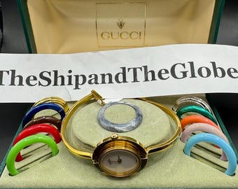 May include: A gold-toned Gucci watch with a white face and a set of colorful interchangeable bezels. The watch is displayed in a green box with the Gucci logo. The box also contains a matching bezel.