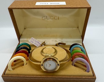 May include: A gold-toned Gucci watch with a white face and interchangeable colored bezels, presented in a wooden box. The box is lined with a tan fabric and features the Gucci logo. The watch has a new battery as of January 2020.